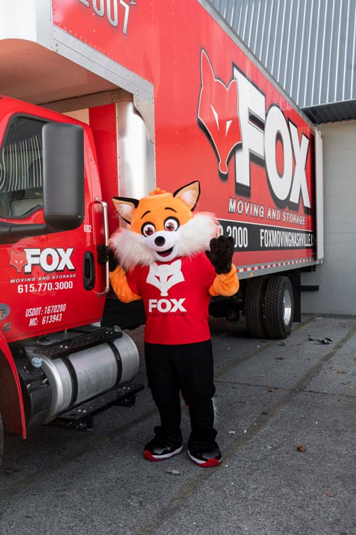 A mascot of Fox Moving and Storage, an orange fox, is standing beside a moving truck
