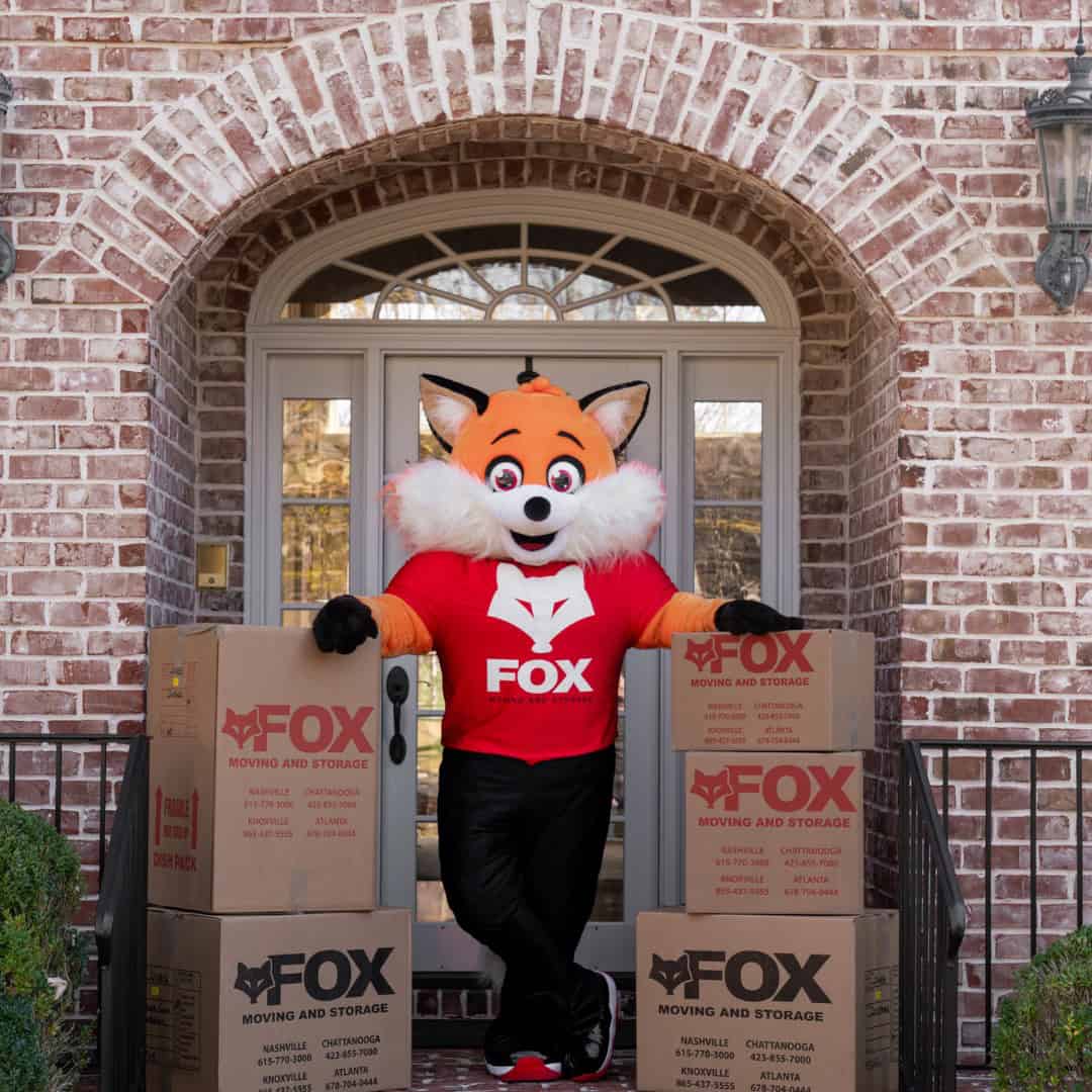 a mascot of fox moving and storage, standing in front of a house, with the moving boxes