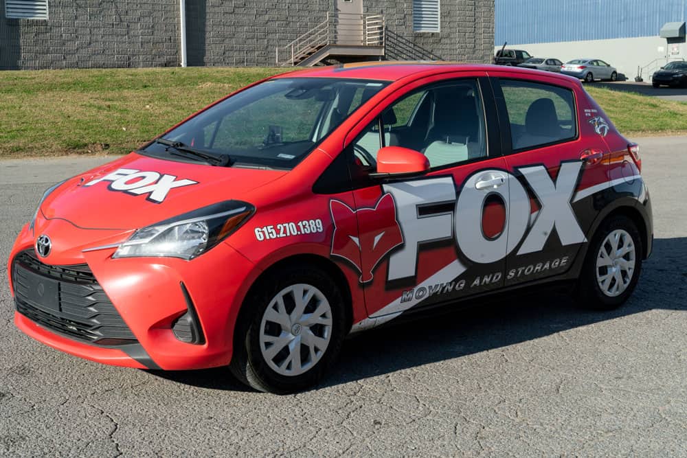 a small red toyota car branded in fox moving and storage's colors standing in front of a storage facility