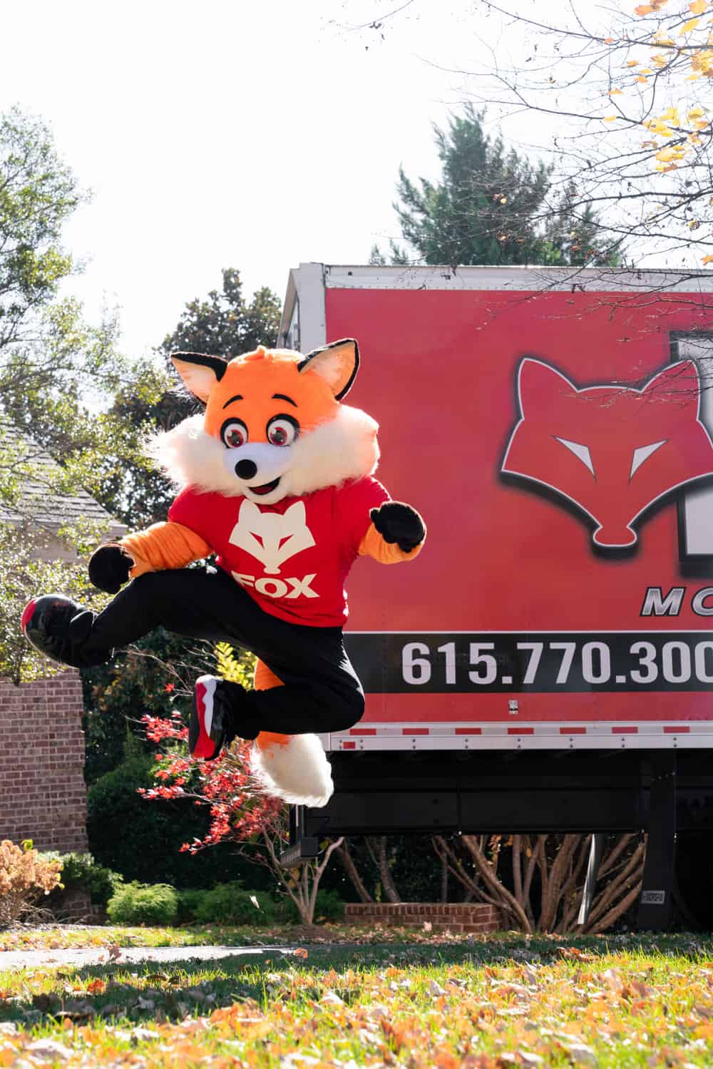an organge fox, a mascot of the fox moving and storage, is jumping up in the air, with the moving truck of fox moving and storage standing in the back.