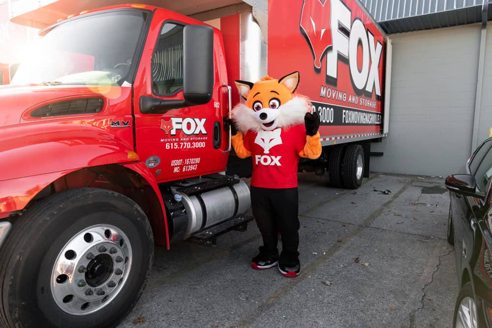 a mascot of fox moving and storage, smiling while standing next to their red moving truck