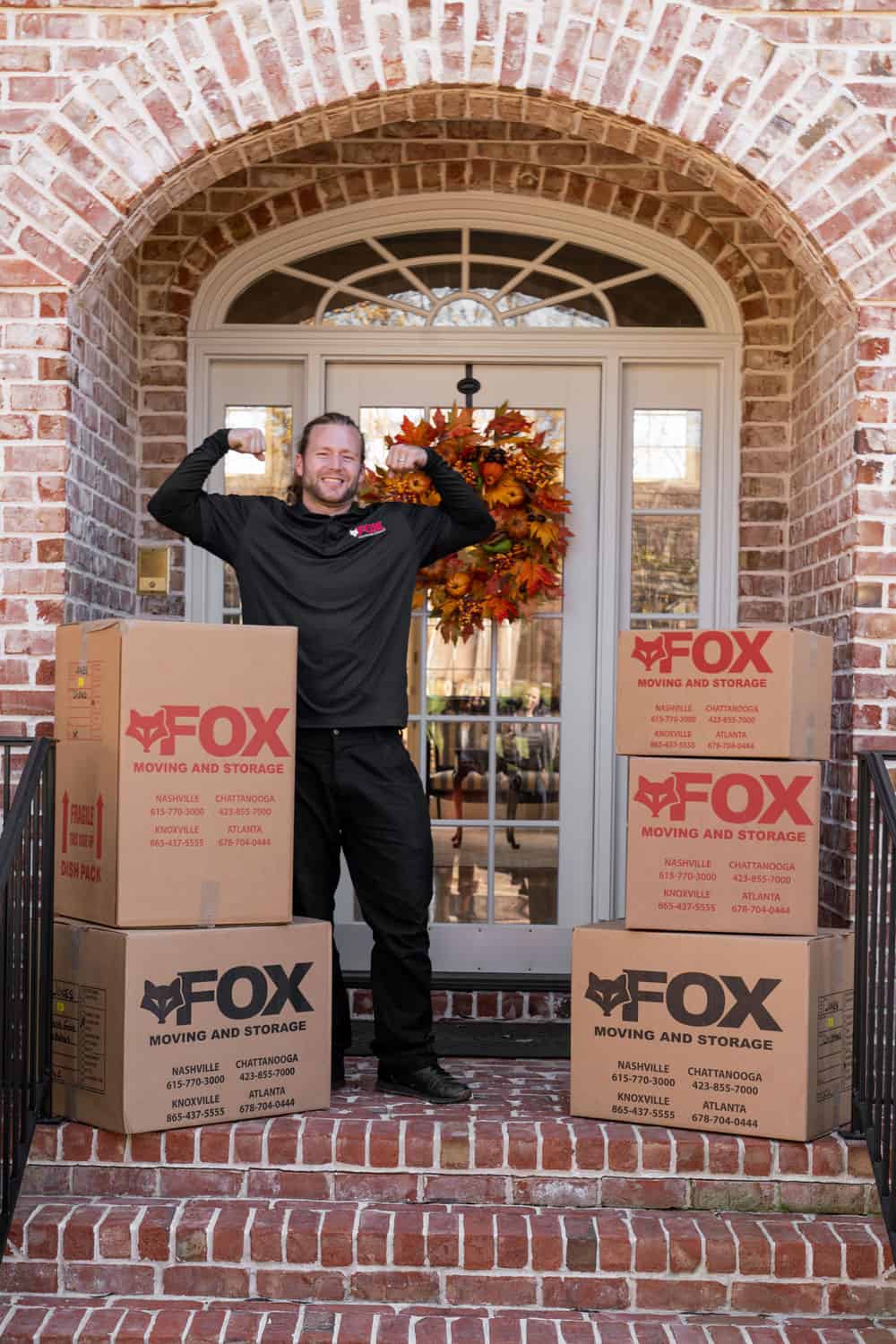 a mover of fox moving and storage, flexing his muscles, while smiling and standing in front of an old house among moving boxes.
