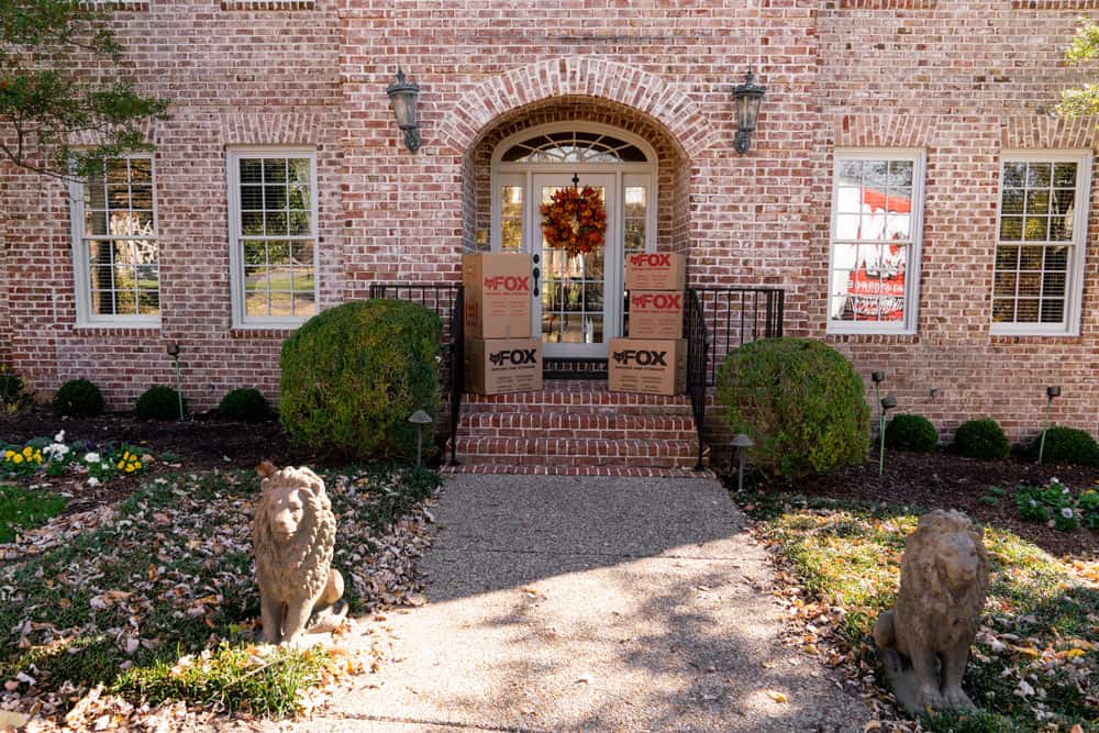 In the picture, we see moving boxes placed on the patio of an old luxurious house, with the brick walls, having large windows and two monuments of lions in front